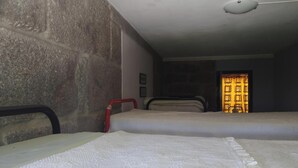 Ein Zimmer mit Steinwand, zwei Betten mit weißer Bettwäsche, einem roten Sessel und einer Tür mit einem gemusterten Vorhang.