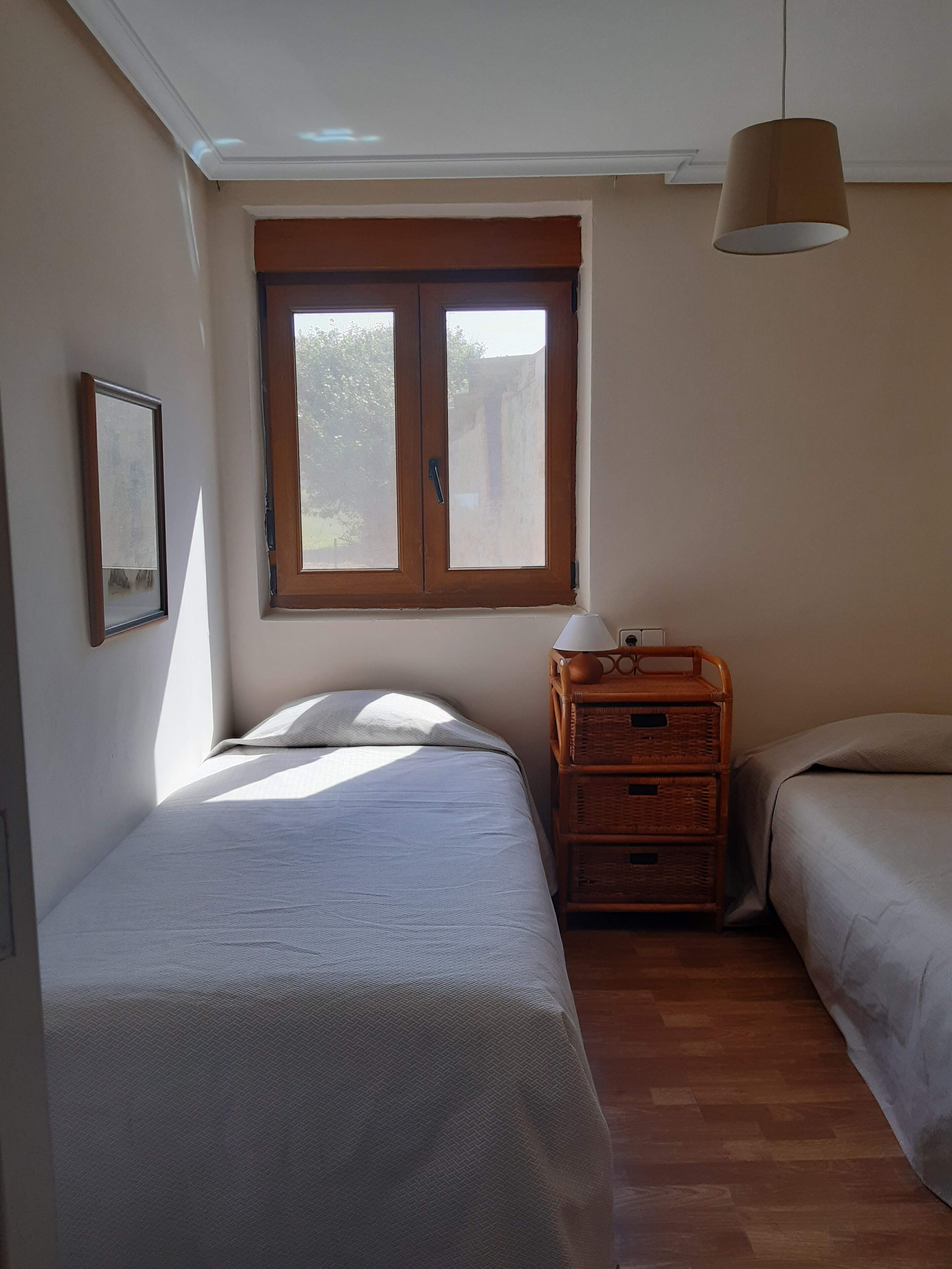 A bedroom with two beds, a window, a hanging lamp, and a wicker storage unit.