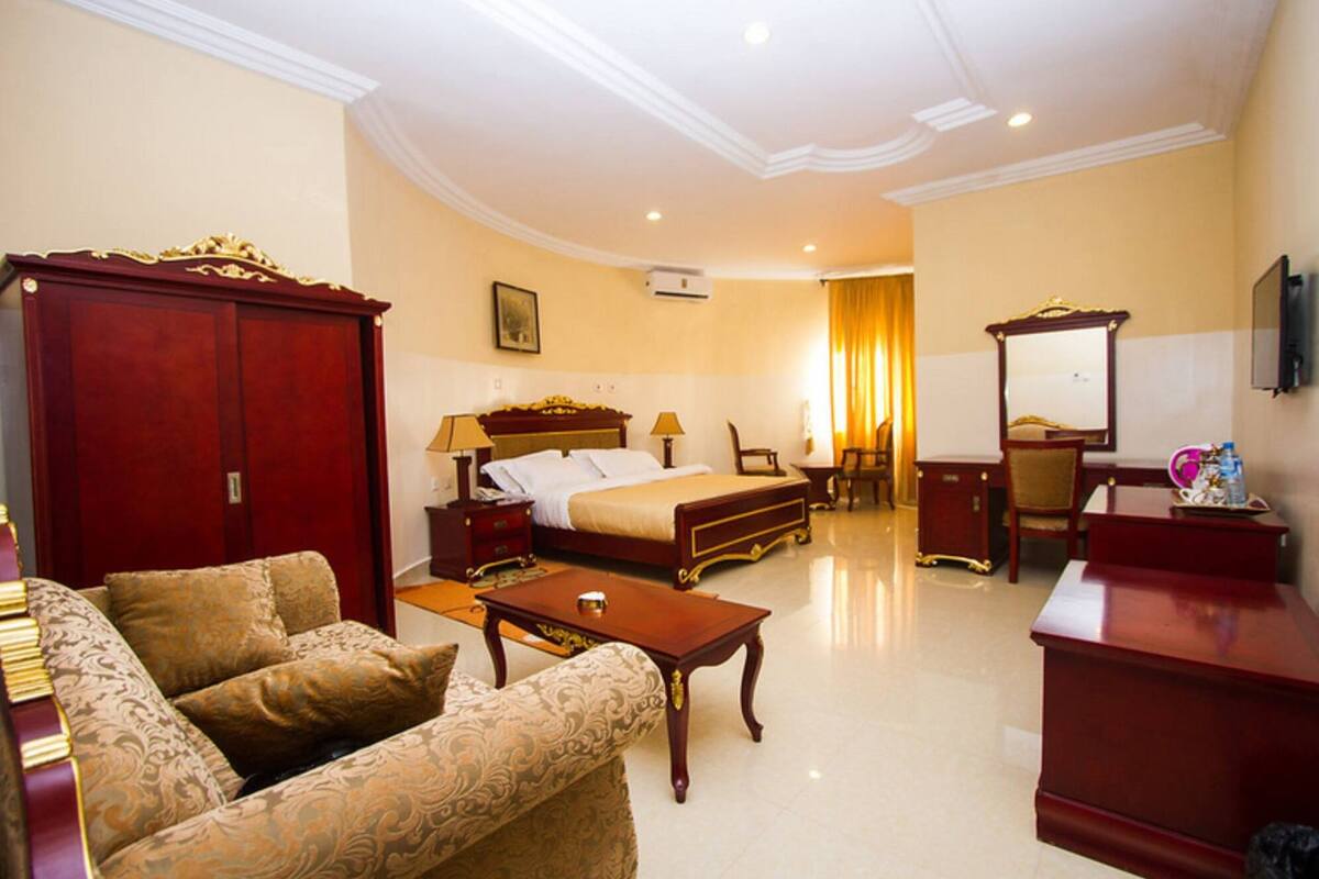 A hotel room with a large bed, a sitting area with a sofa and armchair, a desk with a chair, and a wardrobe.