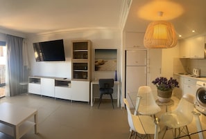 A modern living room with a flat-screen TV, a dining area with a glass table, and a kitchen with a washing machine.