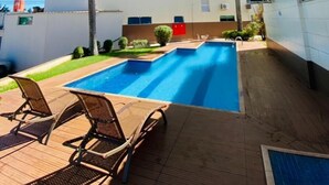Une piscine entourée de chaises longues et bordée de palmiers, avec un immeuble à plusieurs étages doté de climatiseurs.