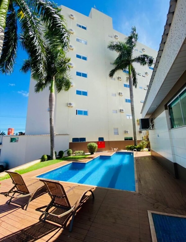 A swimming pool with lounge chairs, surrounded by palm trees and a multi-story building with air conditioning units.