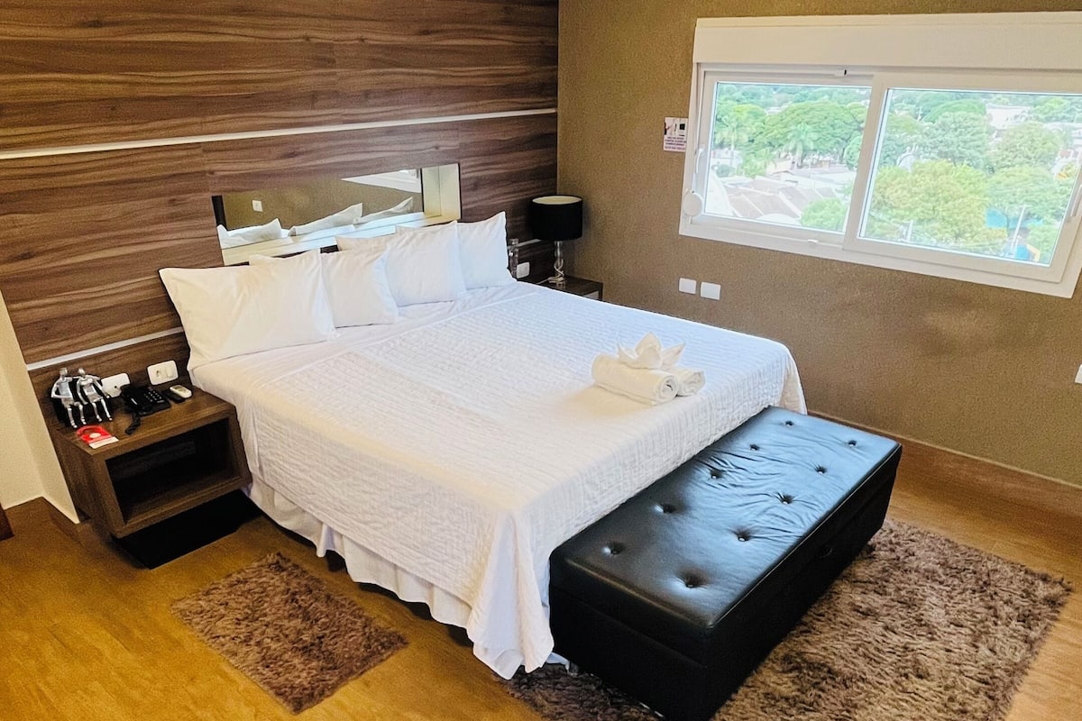 Ein modernes Schlafzimmer mit einem großen Bett, einem Holzkopfteil, Nachttischen, einer Bank und einem Fenster mit Blick.