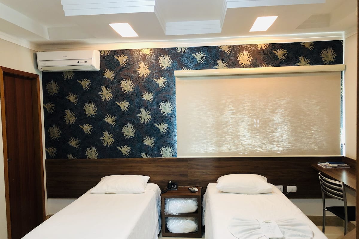 Ein Hotelzimmer mit zwei Einzelbetten, einem Holzkopfteil, einem großen Fenster mit Aussicht, einer eingebauten Klimaanlage und einem kleinen Tisch mit Stuhl.