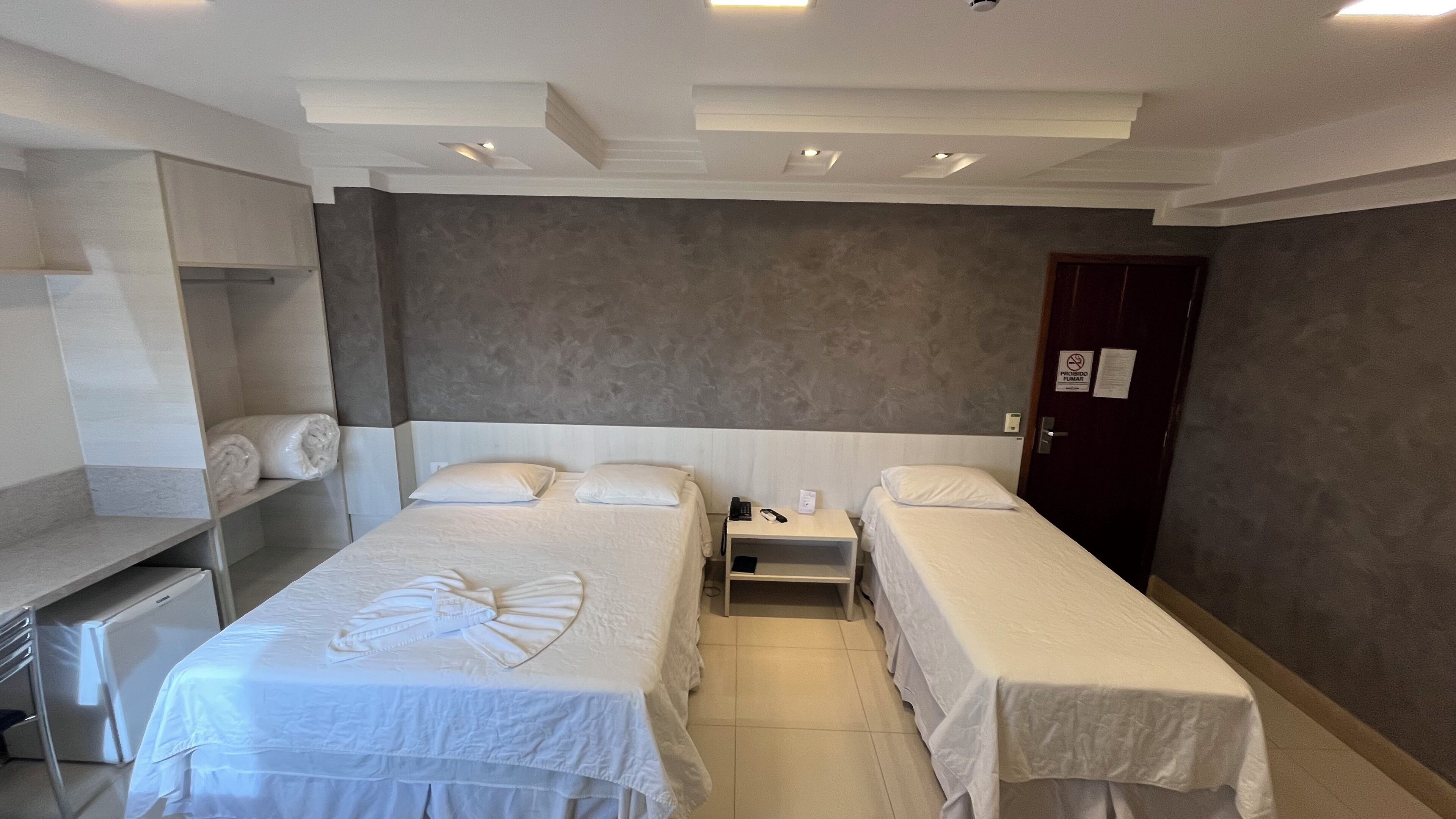 Quarto de hotel com duas camas de solteiro, uma mesa com cadeira, um guarda-roupa e uma porta com placa de 'Proibido Fumar'.
