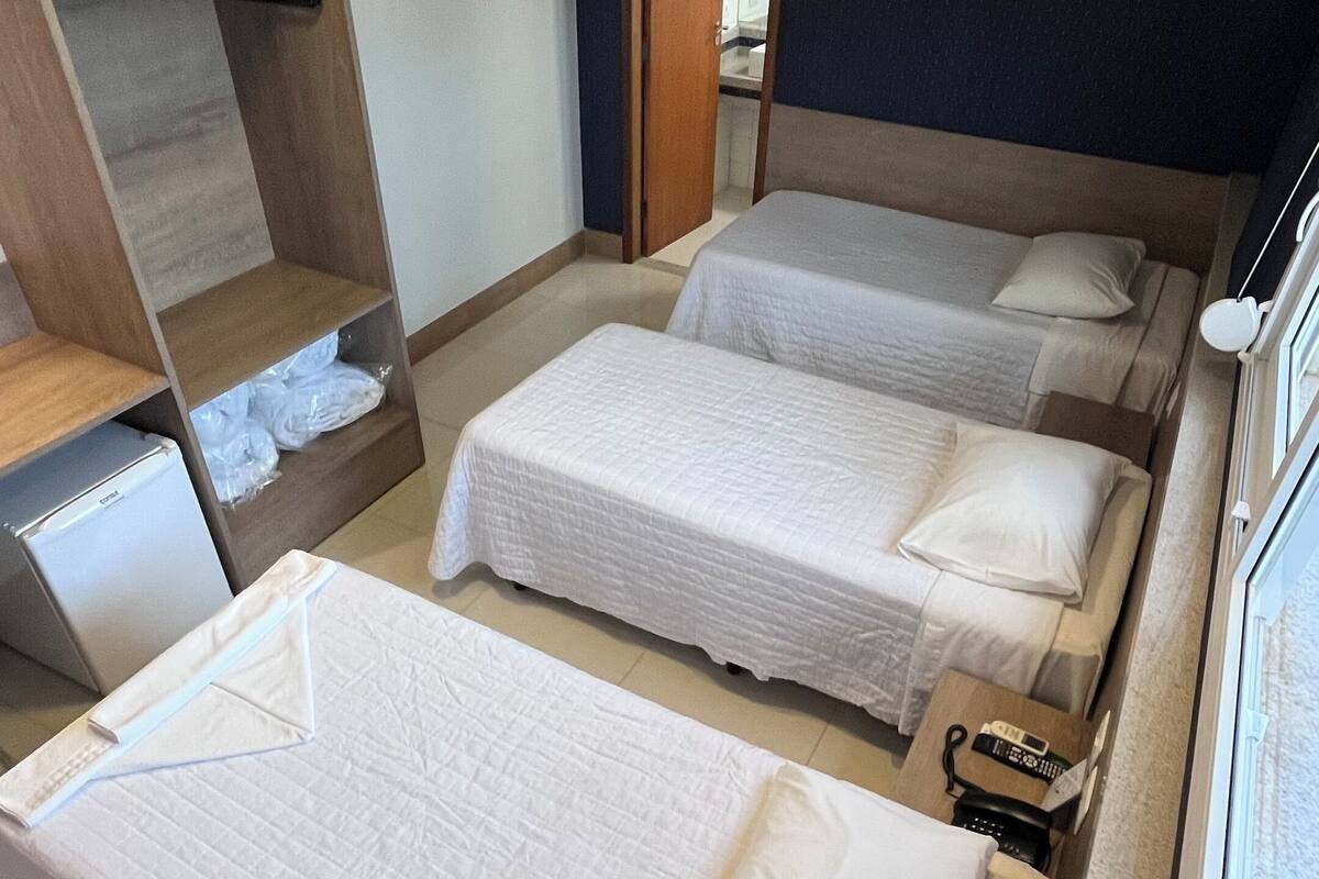 Ein Hotelzimmer mit vier Einzelbetten, einem kleinen Kühlschrank, einem Fernseher und einem durchgegangenen Tür sichtbaren Badezimmer.