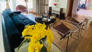 Un salon moderne avec un canapé bleu, une table basse en verre ornée de fleurs jaunes, un téléviseur fixé au mur et un coin repas avec des chaises en métal.