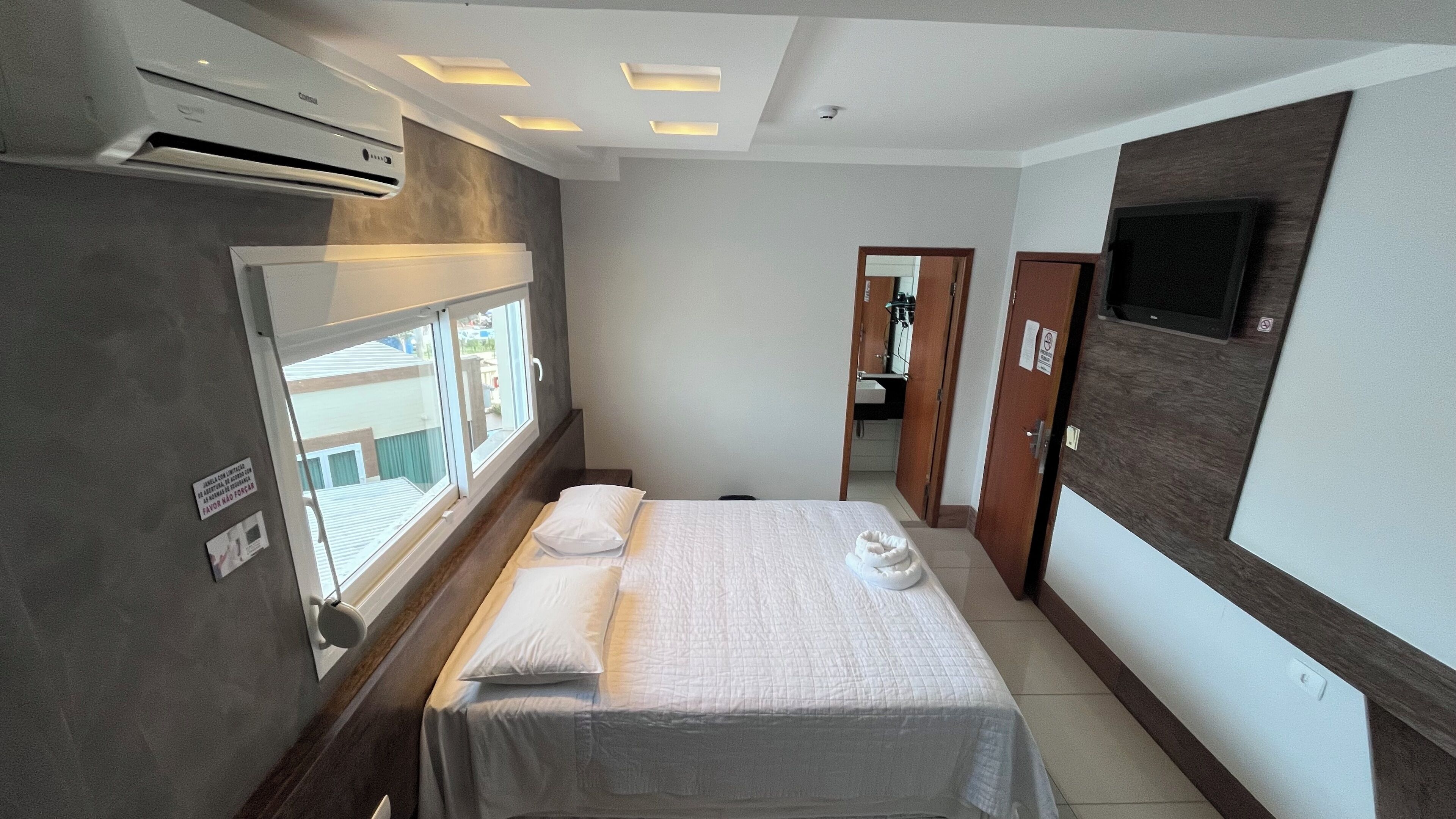Habitación de hotel con cama, ventana, unidad de aire acondicionado, televisor montado en la pared y puerta que da a un baño.