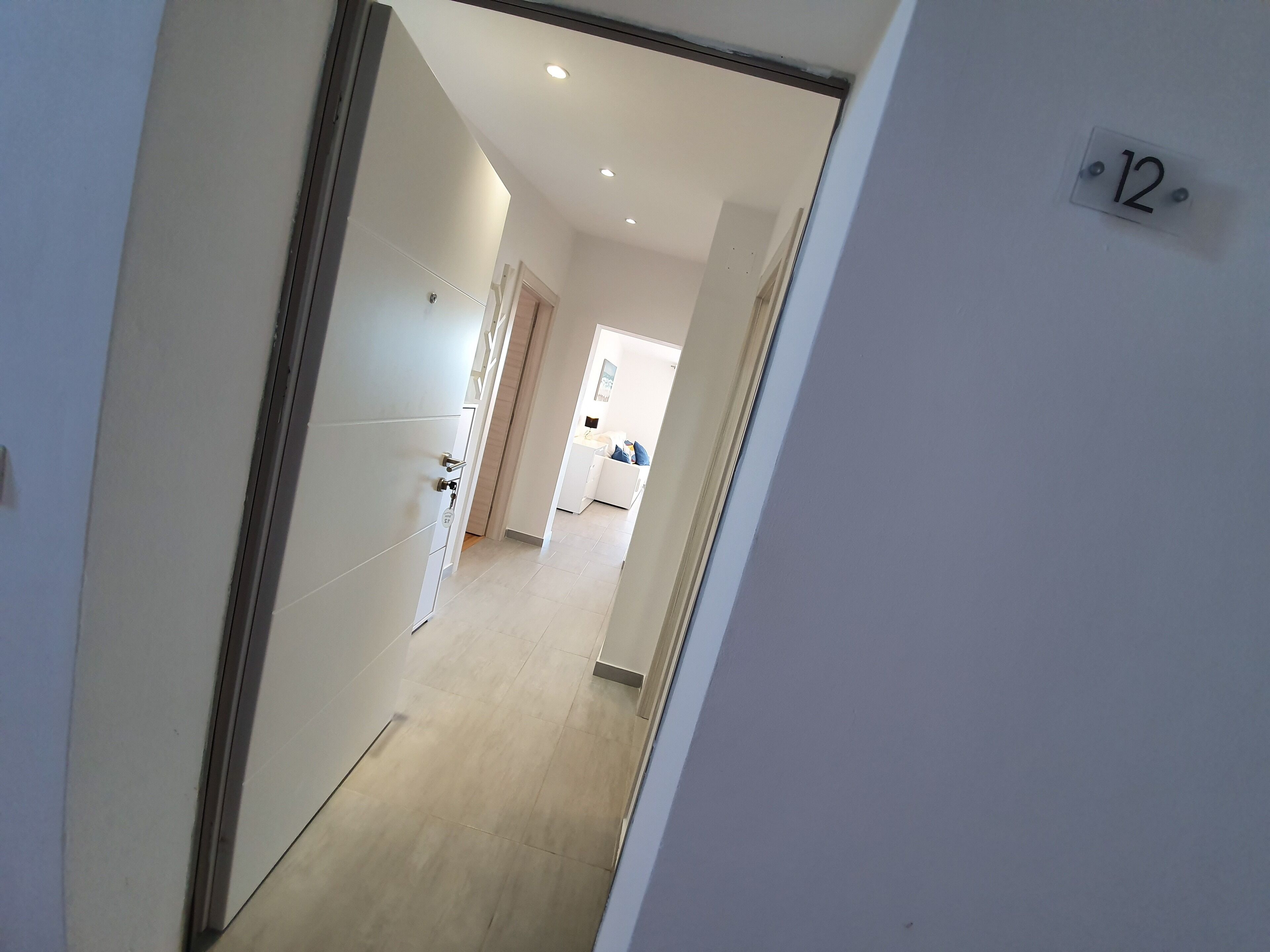 A corridor with a door leading to a room, a number '12' sign on the wall, and a glimpse of a kitchen area.