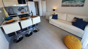 A modern kitchen with white cabinets, a dining area with a white table and orange plates, a white sofa with blue and yellow cushions, and a knitted pouf. - Beach front apartment for family vacation in Mandre (Mandre)