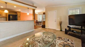 A living room with a glass-top coffee table, a flat-screen TV, and a dining area in the background. - Gardens, St. Mary's, JFK, Good Samaritan, VA (West Palm Beach)