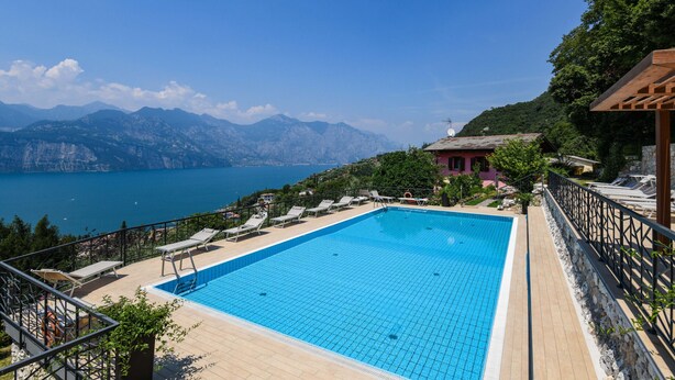 A swimming pool with lounge chairs, surrounded by a wooden deck and a stone wall, overlooking a lake and mountains. - Garden Residence Malcesine Lake View  App. G1 (Malcesine)
