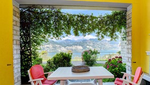 A patio with a table and chairs, a view of mountains and a body of water, and a yellow wall. - Garden Residence Malcesine Lake View  App. G1 (Malcesine)