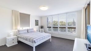 A bedroom with a bed, bedside tables, a desk, a fan, and a window with blinds.