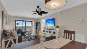 Una sala de estar moderna con ventilador de techo, una mesa de comedor con velas y una televisión grande mostrando una escena de playa.