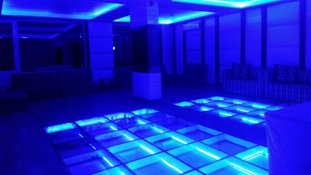 Quarto com chão iluminado em azul e área de estar com sofá e poltronas.