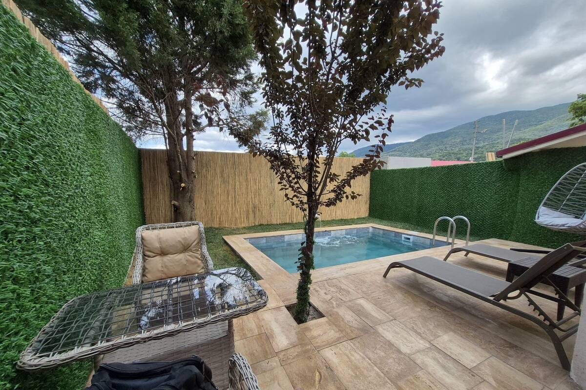 A backyard with a swimming pool, a tree, and outdoor furniture including a chair and a lounge chair.