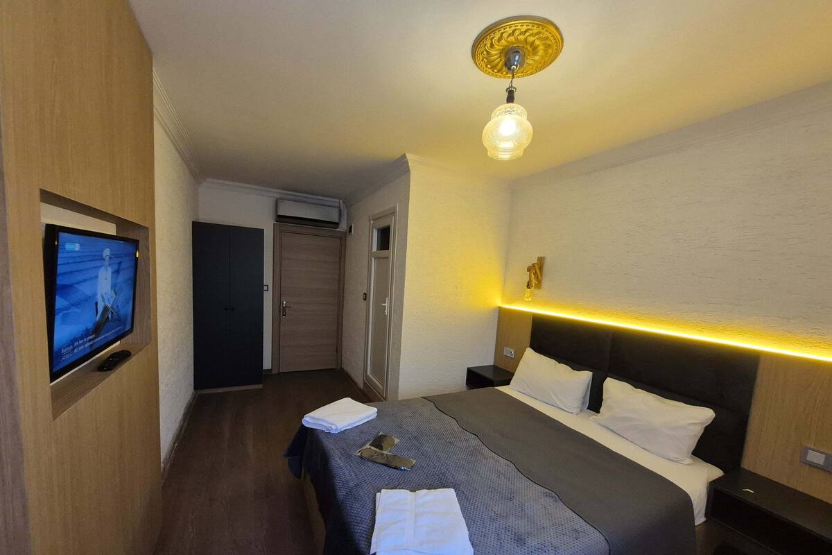 A hotel room with a bed, a TV mounted on the wall, a bedside table, and a ceiling light fixture.