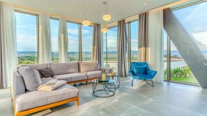 Ein modernes Wohnzimmer mit einem grauen Sofa, einem blauen Sessel und einem runden Couchtisch. Große Fenster bieten Blick auf die Stadtlandschaft.