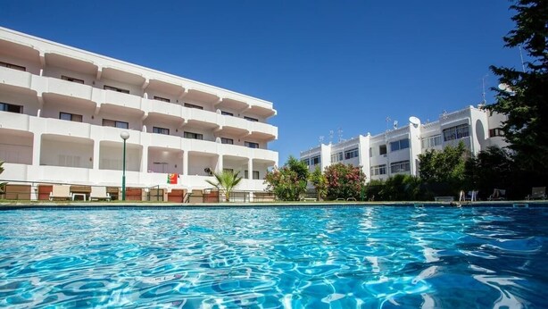 A swimming pool with clear blue water, surrounded by a modern building with multiple balconies and a flag.
