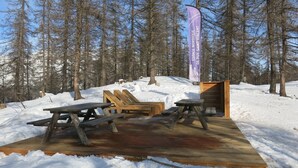 Puinen terassi, jossa on piknikpöytiä ja -tuoleja, ja jonka ympärillä on lunta ja puita.