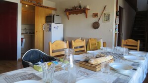 Ruokailutila, jossa on neljälle katettu ruokapöytä, puiset tuolit, jääkaappi ja taustalla keittiö.