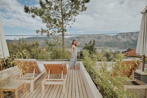 A wooden deck with lounge chairs, a person standing, and a mountainous landscape in the background.