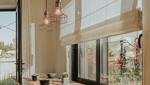A dining area with a wooden table, wicker chairs, and a view of a garden through large windows. - Elsa Modern Apartments. 2 Bed with Jacuzzi Apt 3 (Pogonia)
