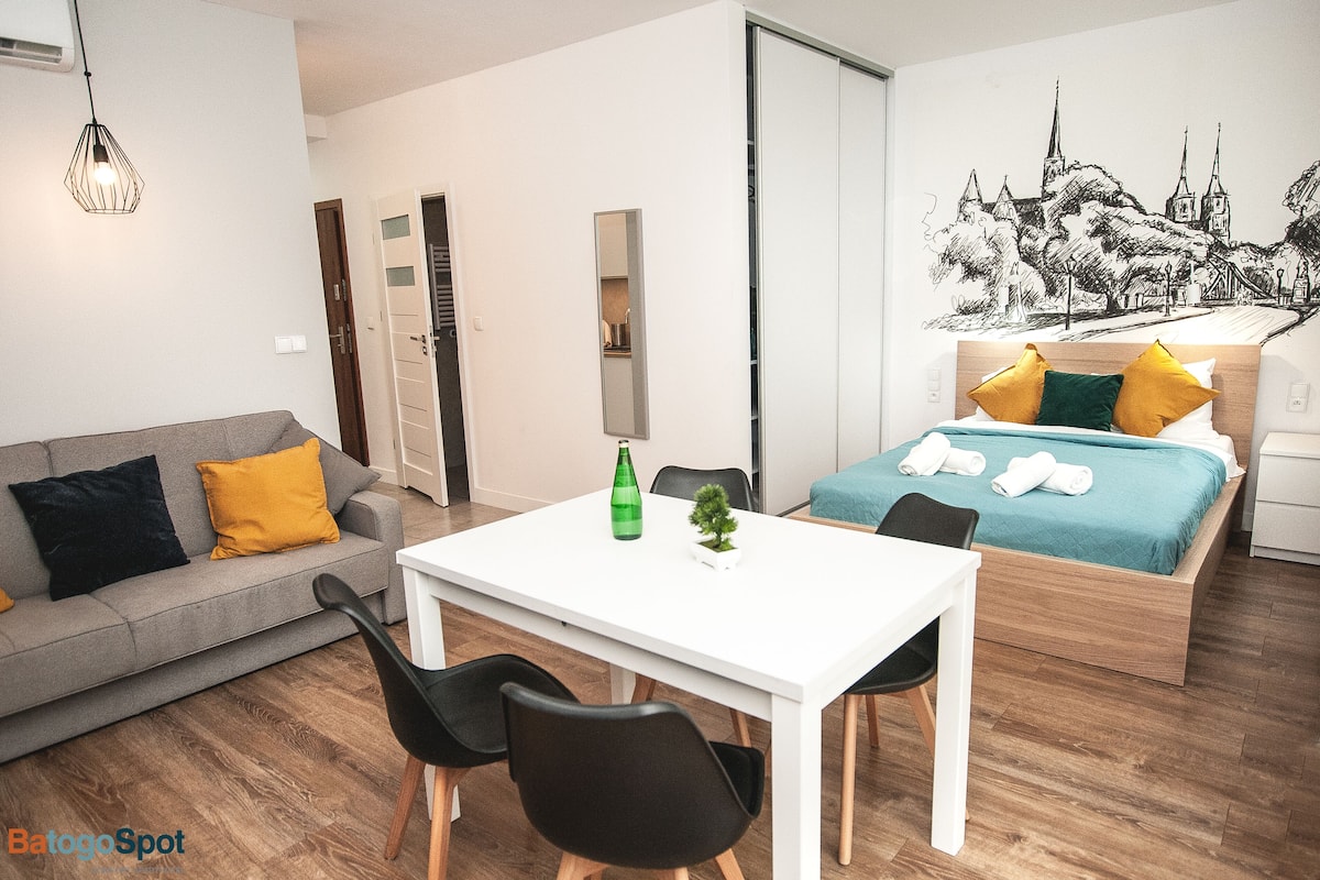 A modern hotel room with a bed, a sofa, a dining table, and chairs. There is a mirror on the wall and a small plant on the table.