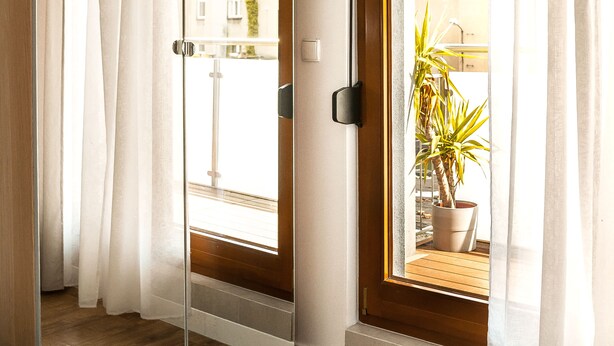 A room with a large mirror, wooden flooring, and a window with a view of buildings and a potted plant. - BatogoSpot Tumski (Wroclaw)