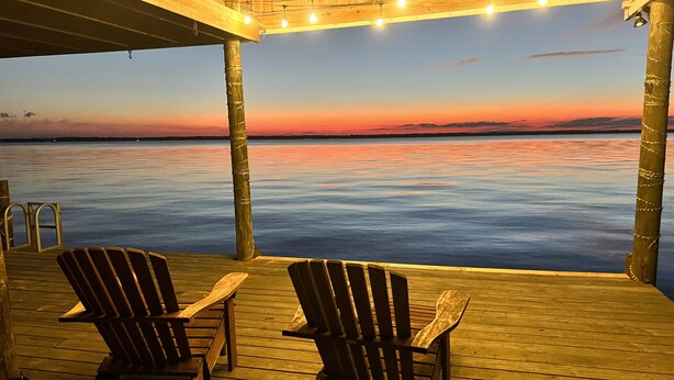 Eine Holzterrasse mit zwei Adirondack-Stühlen, die auf ruhiges Wasser bei Sonnenuntergang ausgerichtet ist.