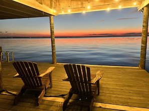Une terrasse en bois avec deux fauteuils Adirondack donnant sur une étendue d'eau calme, au coucher du soleil.