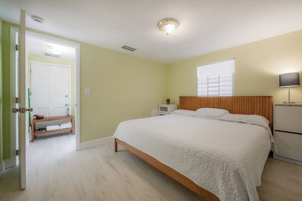 A bedroom with a bed, a nightstand, a lamp, a door, and a shelf with towels.