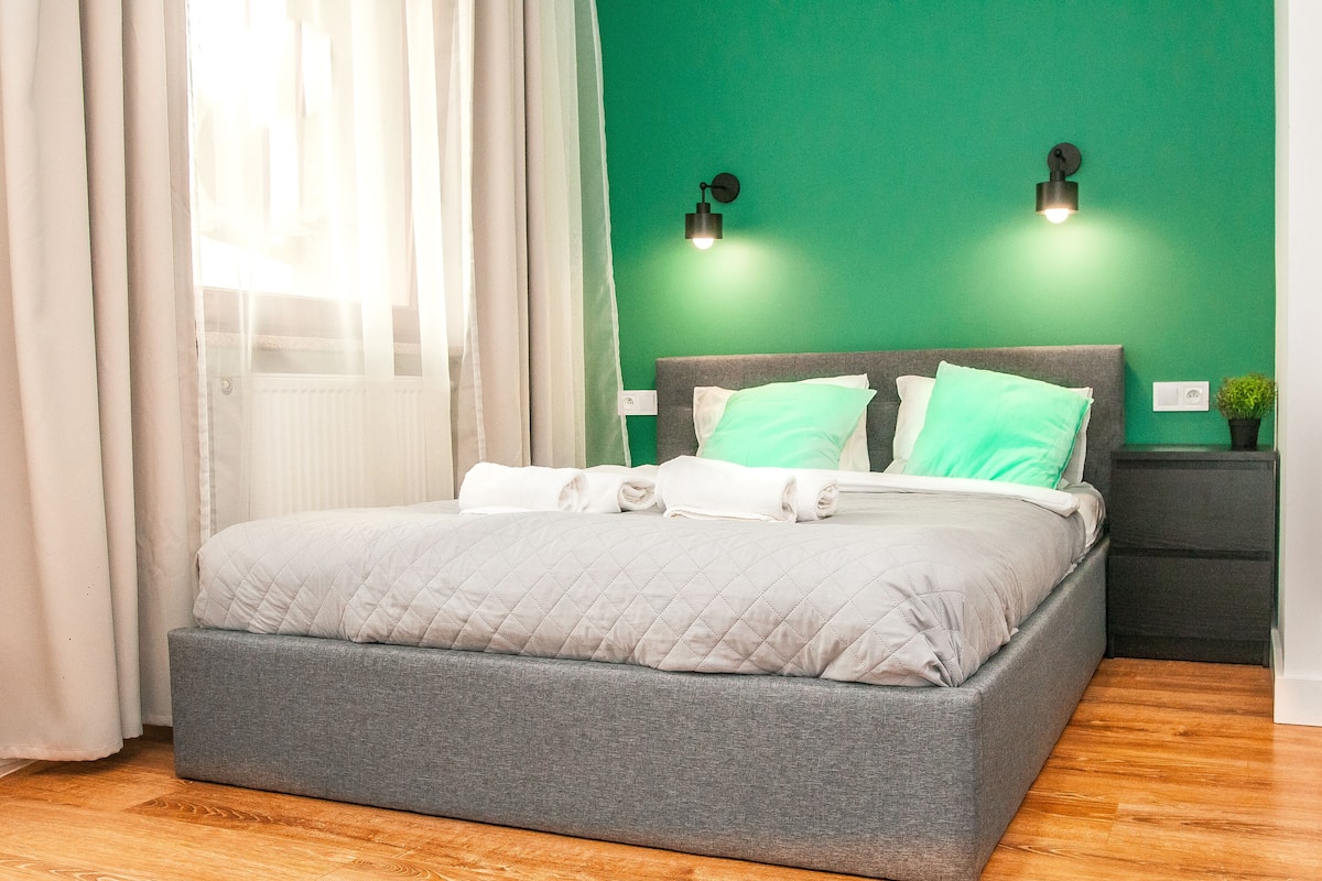 Una cama bien hecha con colcha gris, cabecera tapizada en gris y dos almohadas verdes. Hay dos luces fijadas a la pared, de color verde, una pequeña planta en maceta sobre una mesita de noche y suelo de madera.