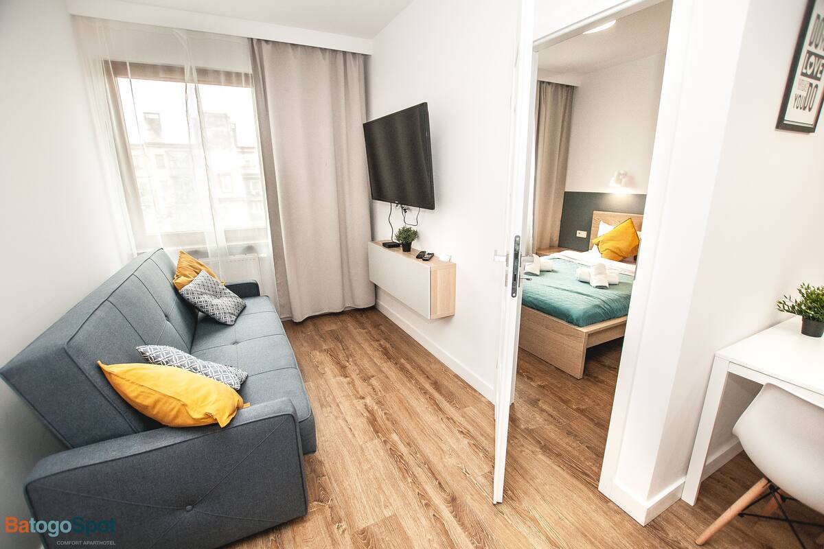 Una habitación de hotel moderna con un sofá gris, una cama con almohadas amarillas, un televisor de pantalla plana y un escritorio.