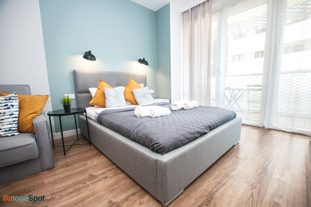 Une chambre moderne avec un lit gris, de nombreux coussins, une petite table avec une plante et une vue sur l’extérieur grâce à de grandes fenêtres.