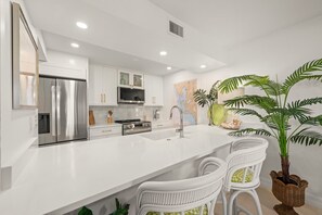 A modern kitchen with white cabinetry, stainless steel appliances, a large island with bar stools, and decorative plants.