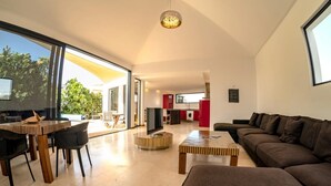 A modern living room with a large window, a dining area, and a kitchen visible through an open door. - Casa Yuna with heated pool in El Roque (El Roque)