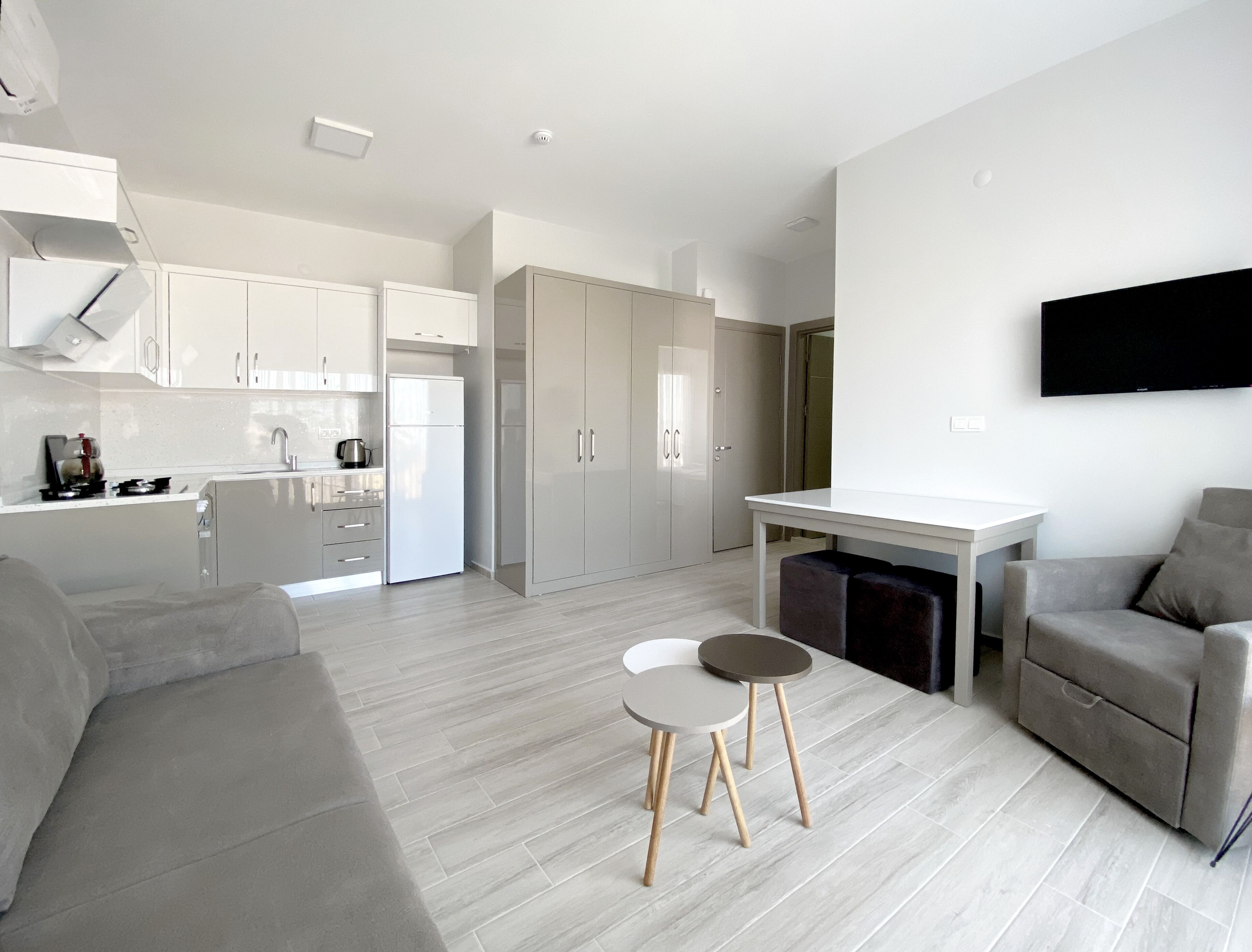 A modern, open-plan living and dining area with a kitchen, grey sofas, a dining table with stools, and a flat-screen TV mounted on the wall.