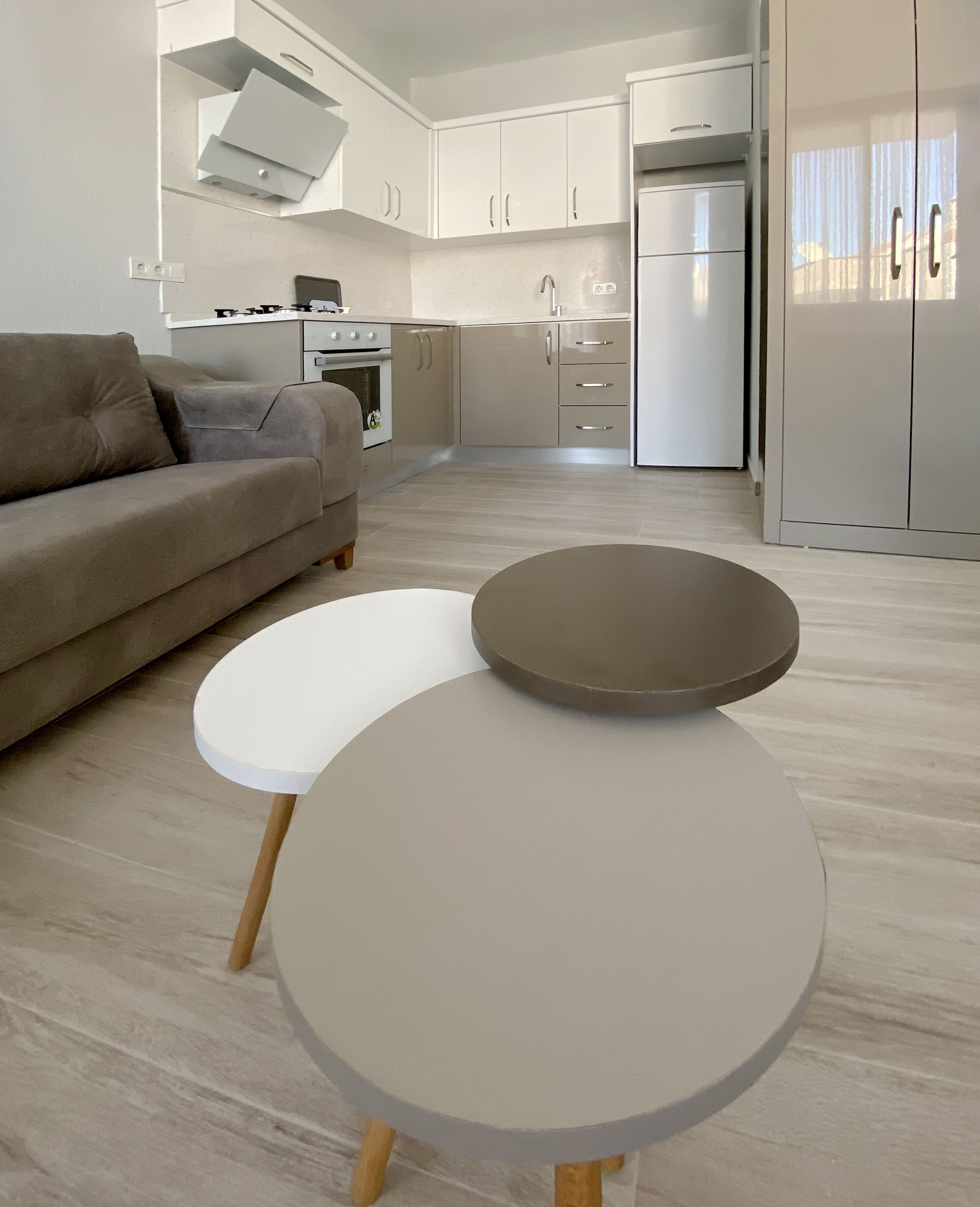 A modern kitchen with white cabinets, a built-in oven, and a refrigerator. There is a beige sofa and two round tables with wooden legs.