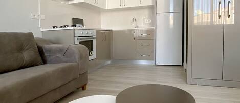 A modern kitchen with white cabinets, a built-in oven, and a refrigerator. There is a beige sofa and two round tables with wooden legs.