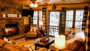 A cozy cabin interior with a fireplace, plush seating, and large windows offering a view of the snowy outdoors. - Riverfront Mountain Cottage Retreat (Estes Park)