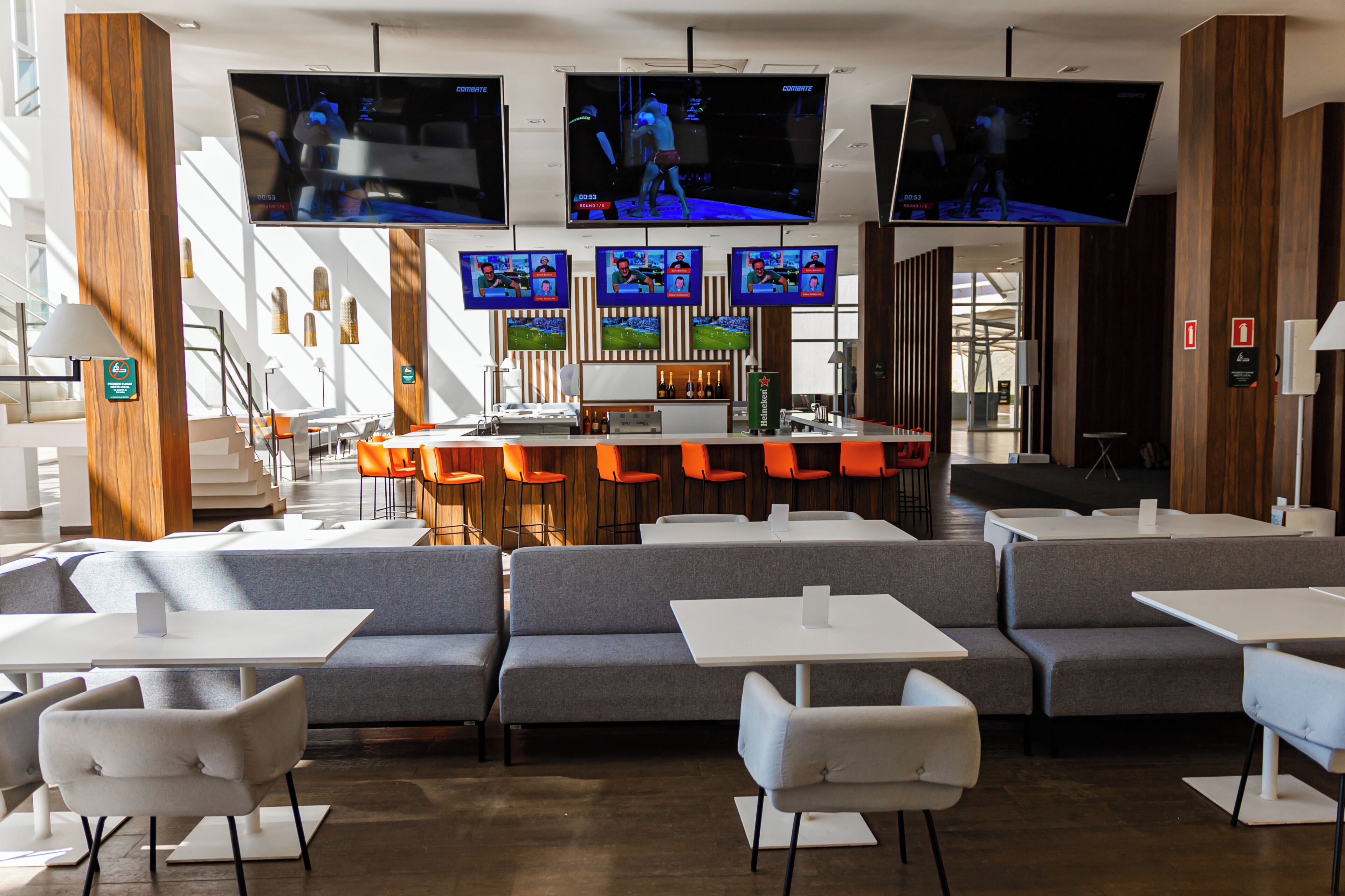 Un área de bar moderna con varias pantallas mostrando contenido deportivo, diferentes disposiciones de asientos y una barra.