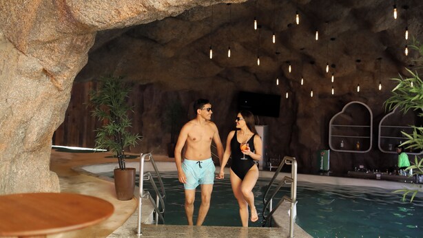 A man and woman walking towards a pool in a cave-like setting with hanging lights and a bar area in the background. - Tauá Resort & Convention Alexânia (Alexania)