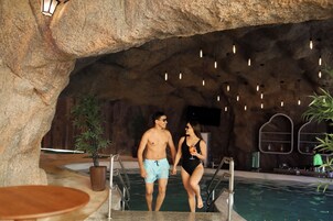 Un hombre y una mujer caminando hacia una piscina en un entorno que recuerda a una cueva, con luces colgantes y un área de bar al fondo.