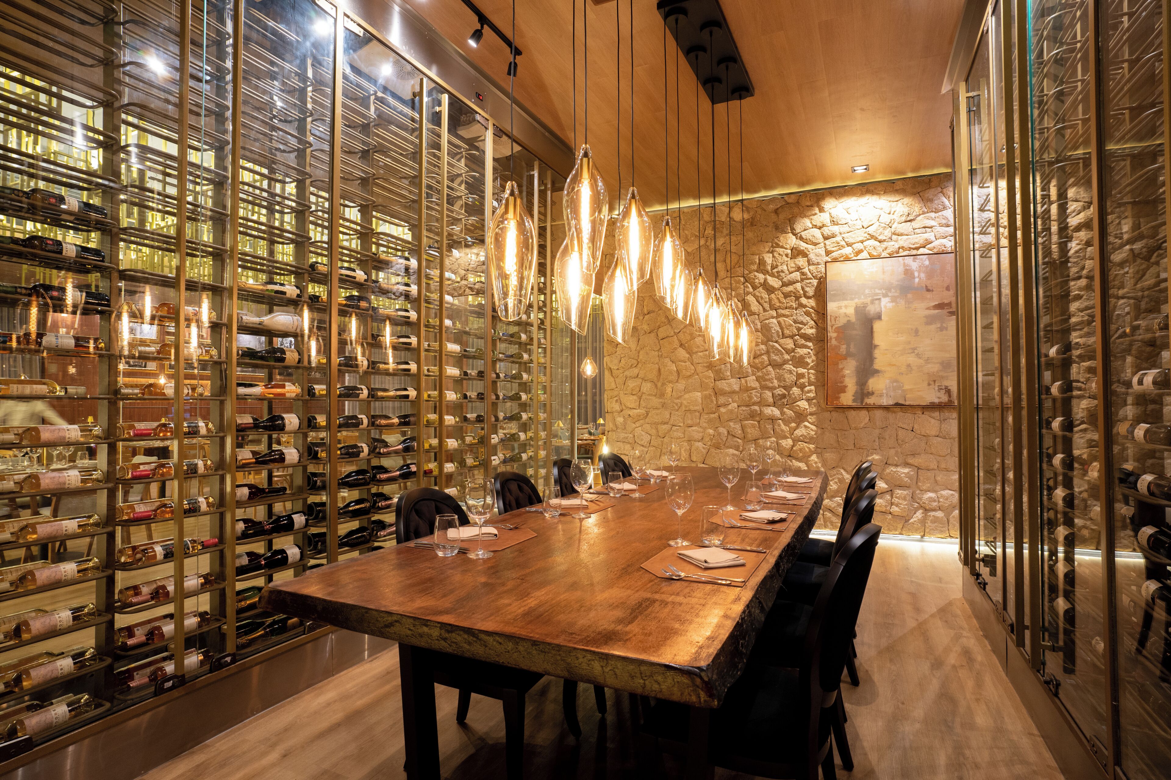 Una bodega con una larga mesa de madera y sillas, rodeada de estantes de vidrio llenos de botellas de vino.
