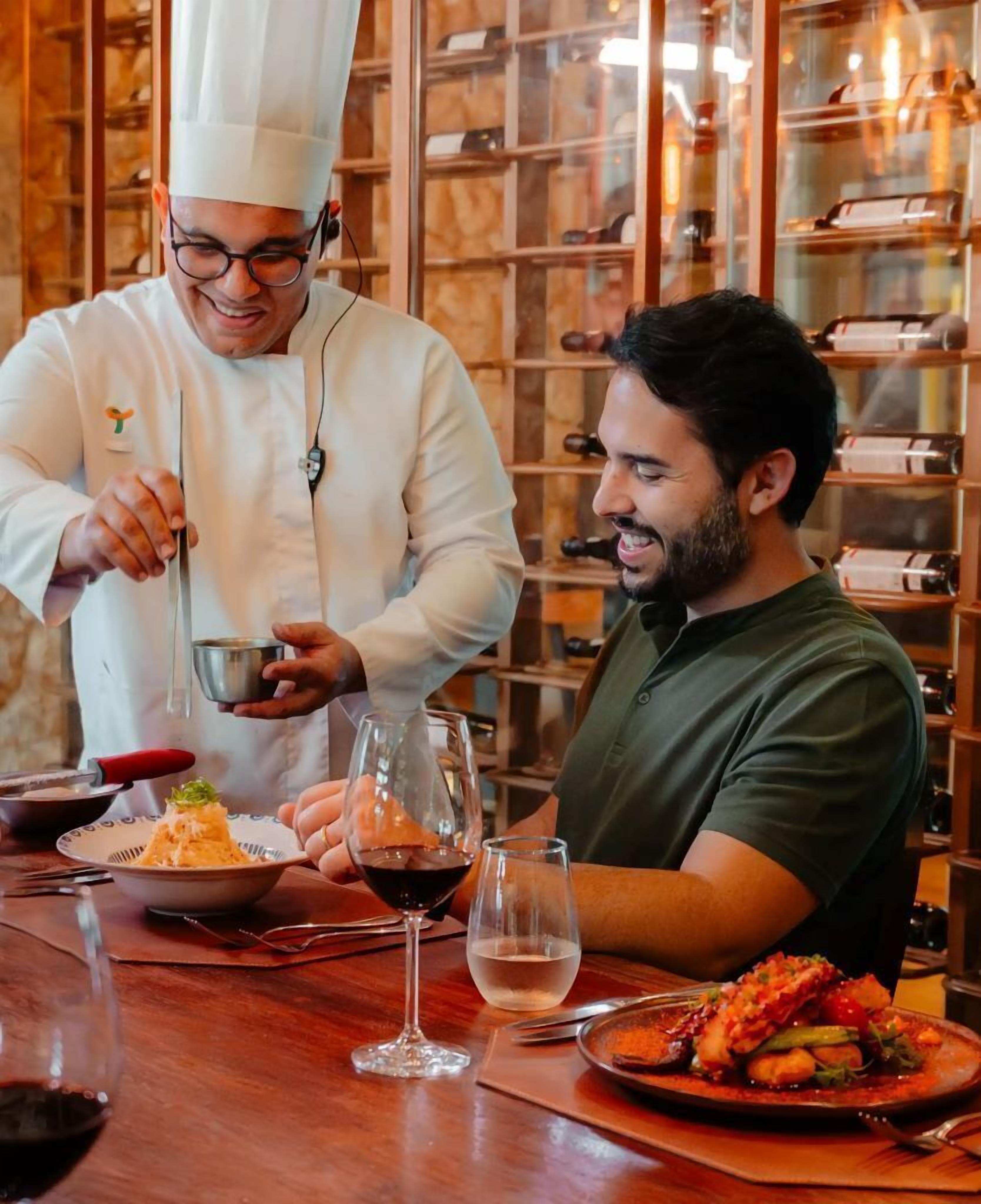 A chef serving food to a guest in a restaurant with a wine rack in the background.