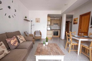 A living room with a sofa, a coffee table, and a dining area with chairs and a table. There is a kitchen visible in the background. - Lanzahost "Casita el Molino" (Costa Teguise)