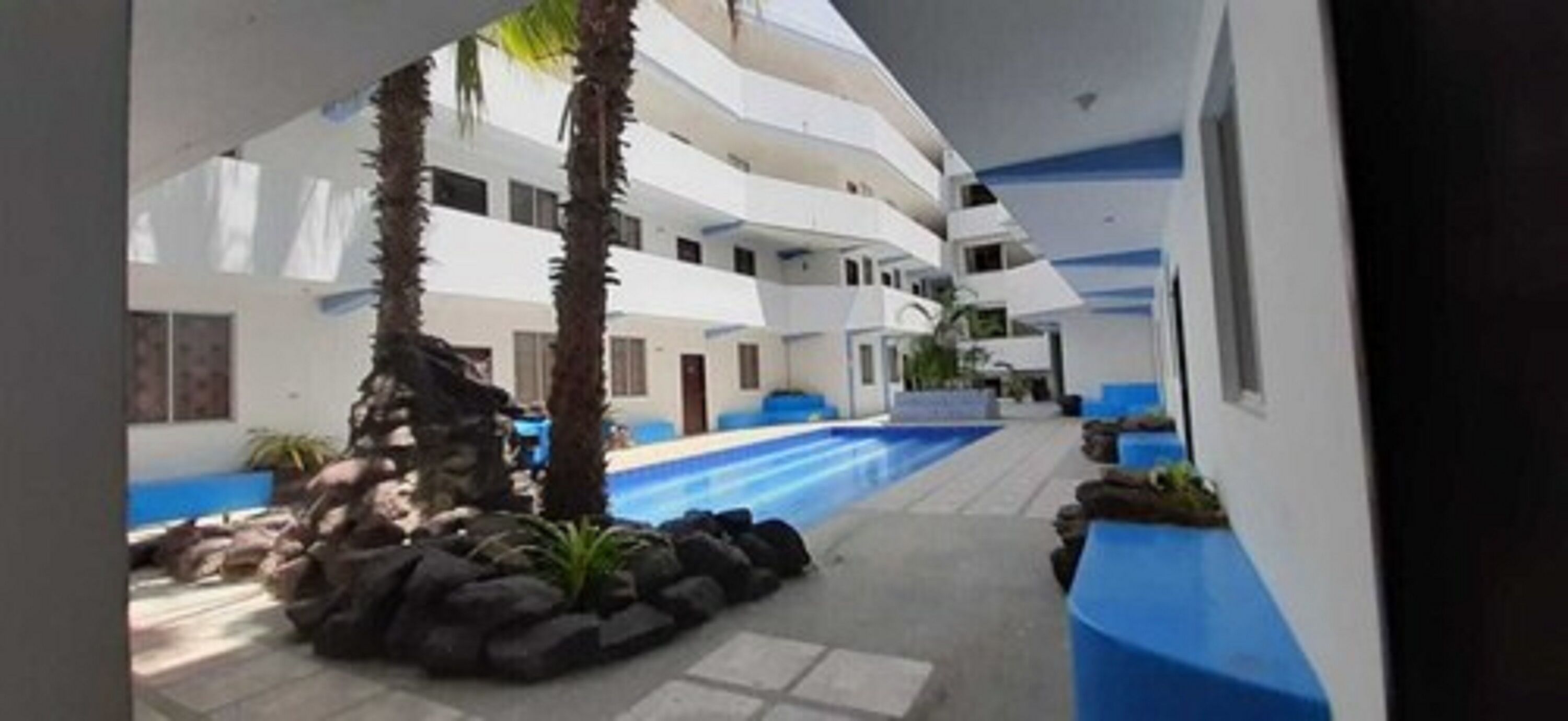 A multi-level hotel with a central swimming pool, surrounded by palm trees and stone features.