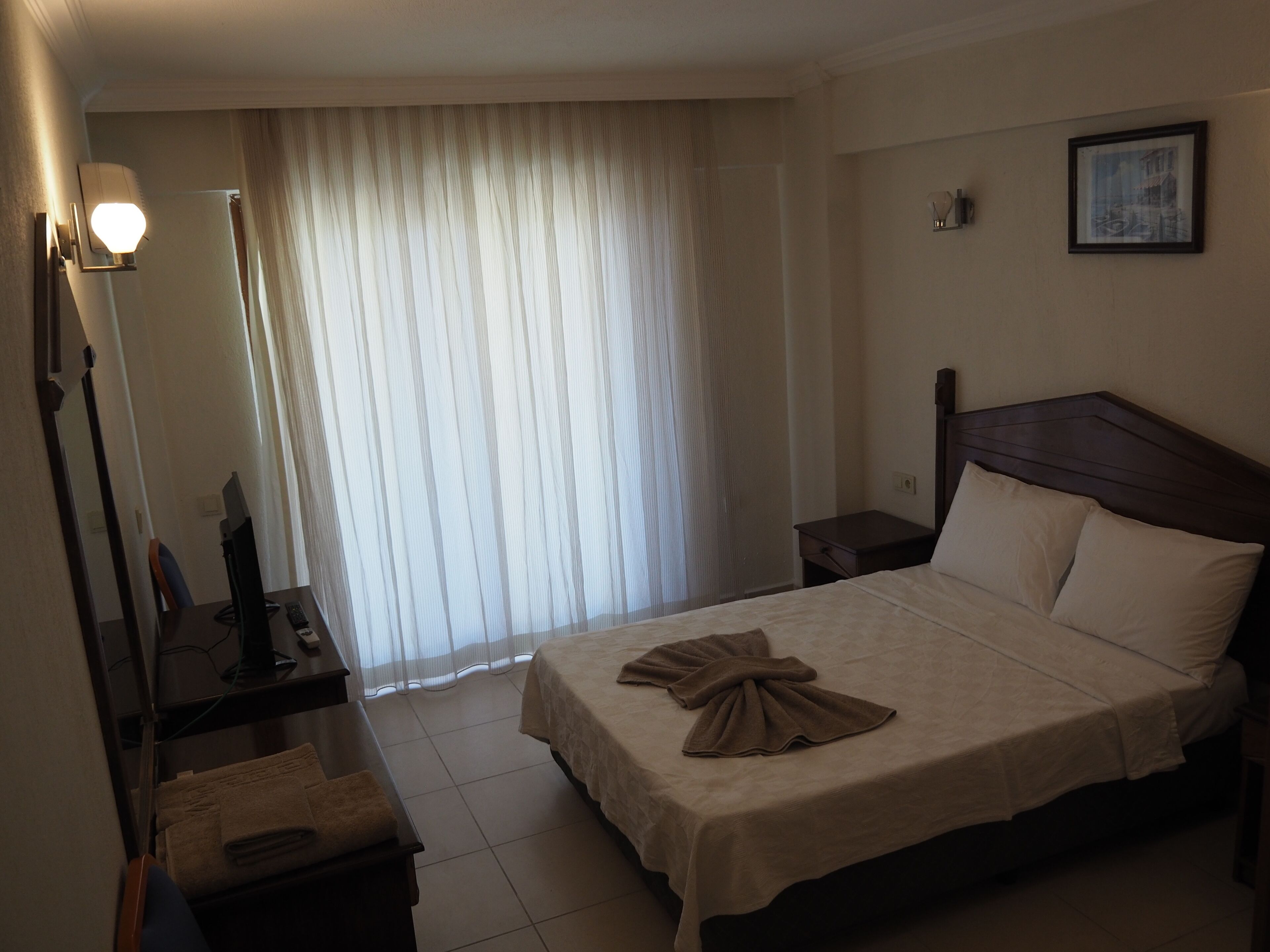 A hotel room with a bed, bedside tables, a desk with a computer, and a window with curtains.